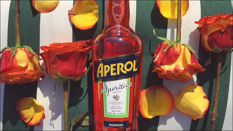 botella de Aperol con rosas naranja sobre superficie de rayas